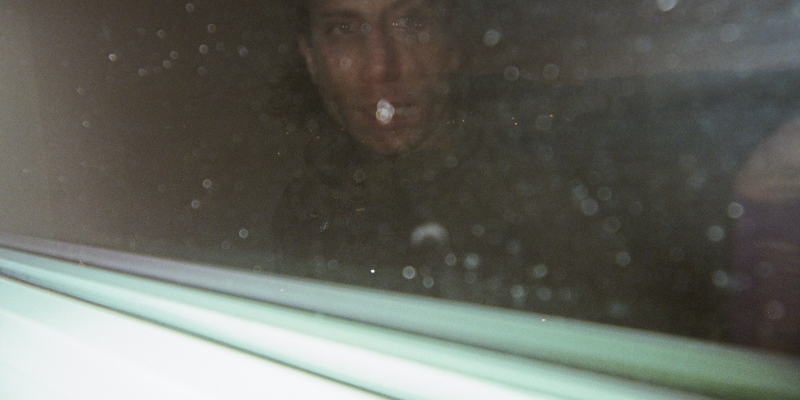 Photo of a man’s face reflected in a dark window. Drops of rain on the glass look like tears on his face. In the bottom left you can see the window pane and the word “Crackdown” written in dark block letters