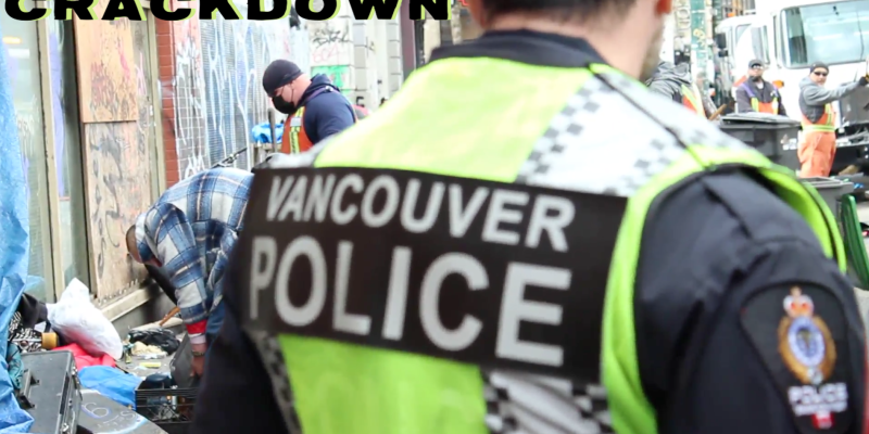 A Vancouver police officer stands in the foreground with his back to the camera. Behind him a man is hunched over grabbing his belongings as city workers clear out the sidewalk area where he was tenting.